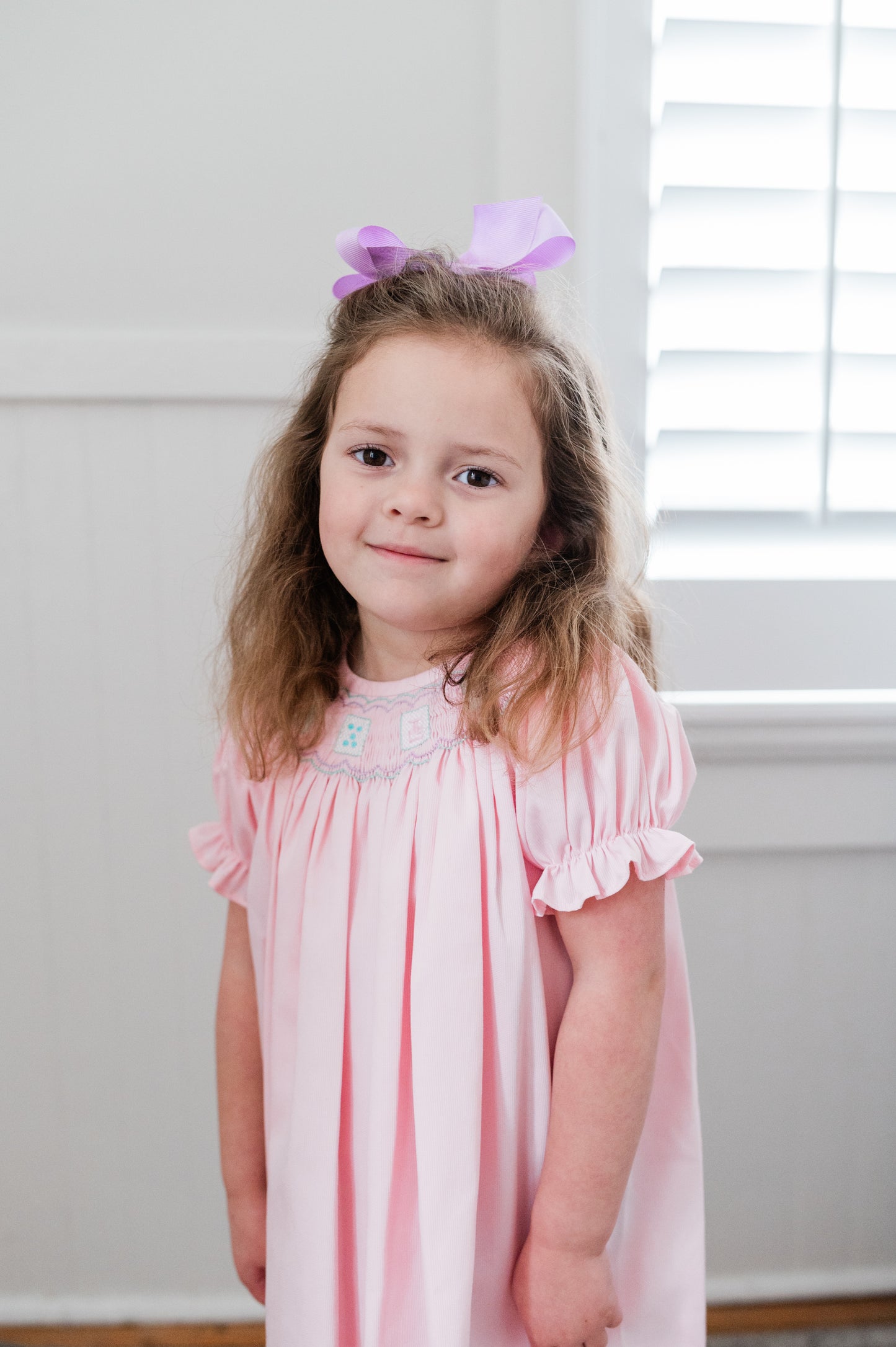 Young girl wearing a pink dress with ruffled sleeves in a room with white walls and a window.