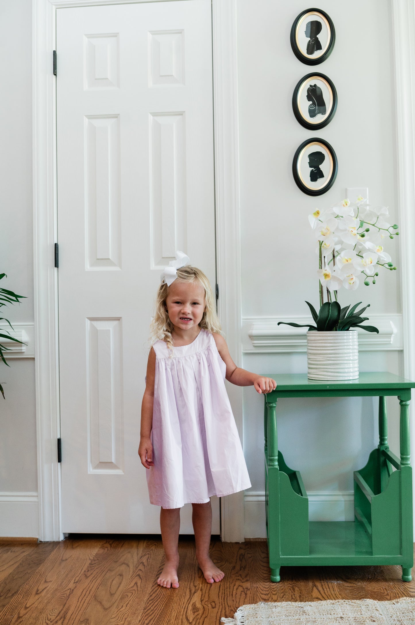 Young girl in a lavender dress standing next to a green table with a plant in a white room.