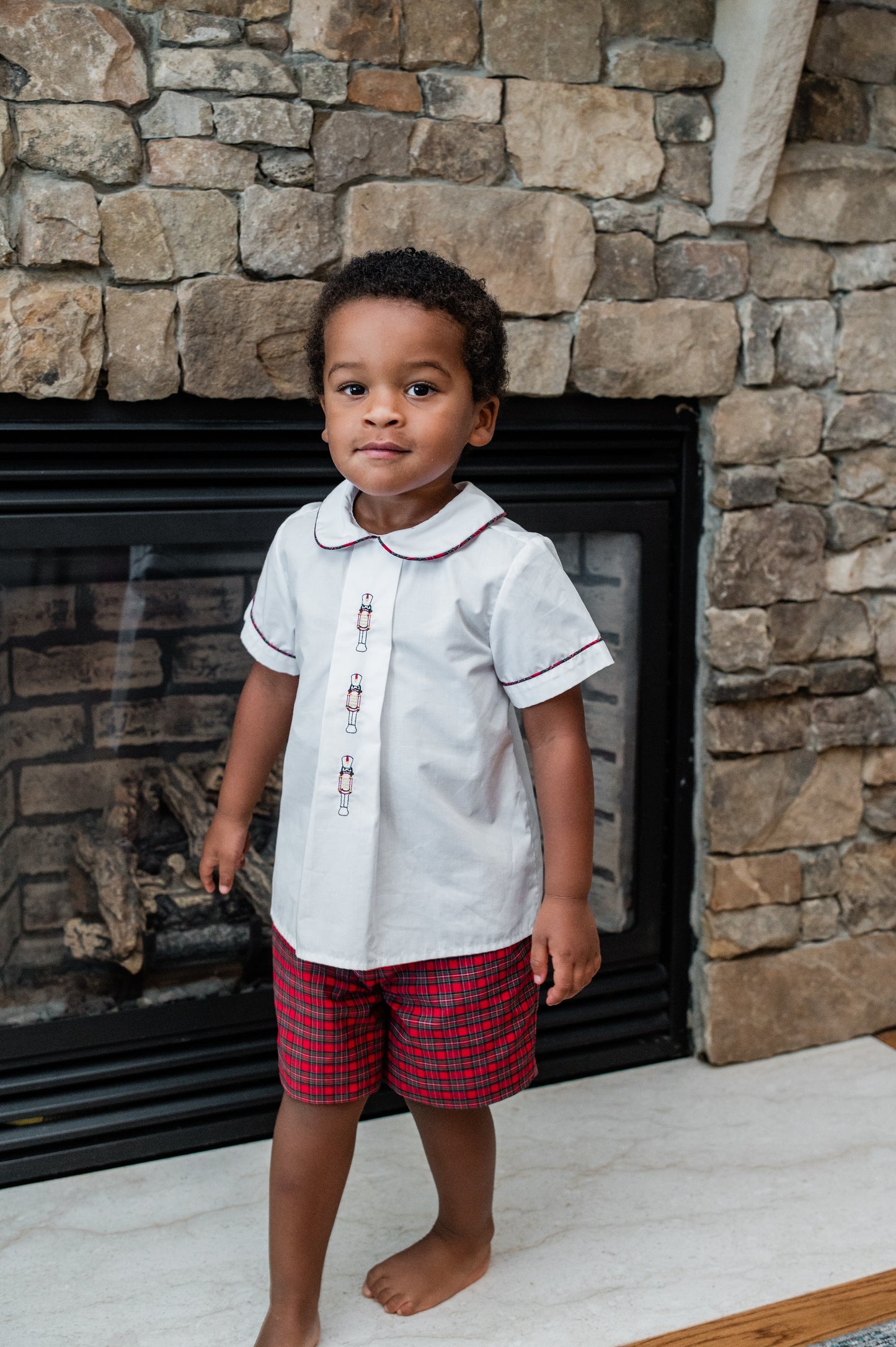 Child wearing a white shirt with red trim and red plaid shorts standing in front of a stone fireplace.
