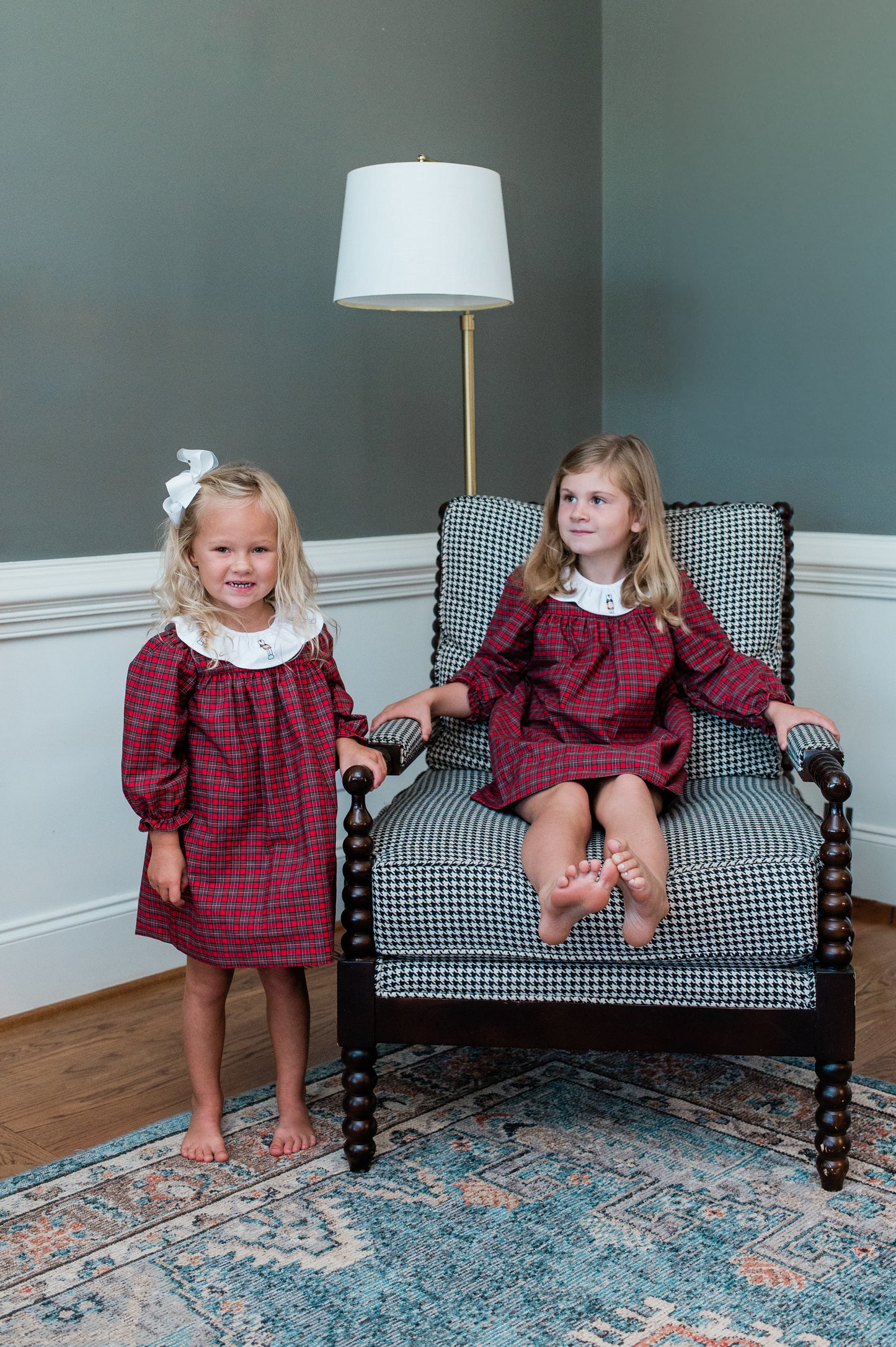 Two young girls in matching red plaid dresses sitting on a checkered chair in a room with a lamp and decorative rug.