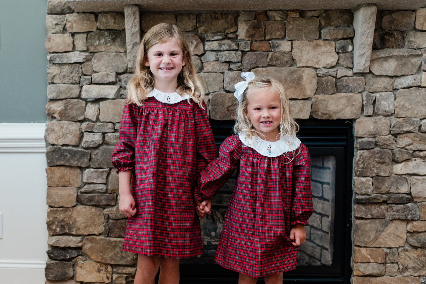 Two young girls in red plaid dresses standing in front of a stone fireplace.