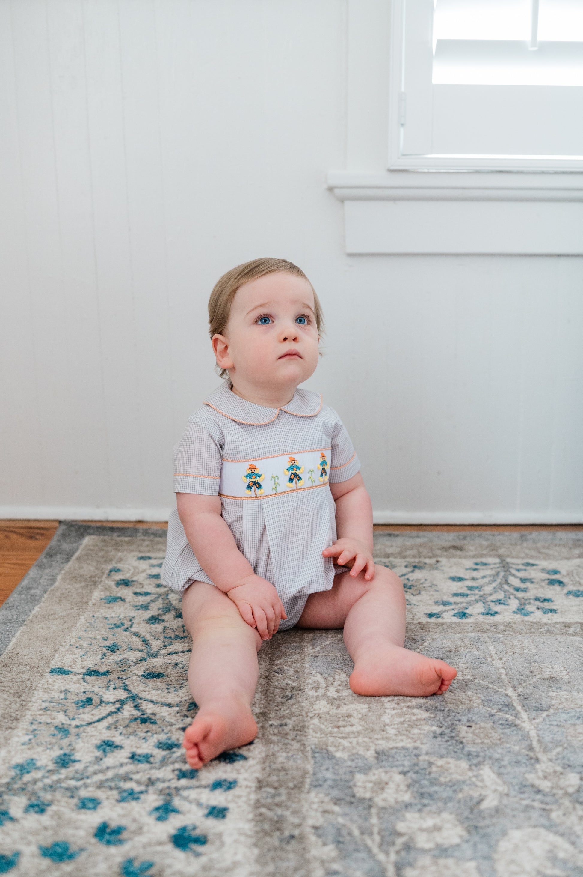 Baby sitting on a patterned rug in a room with white walls.
