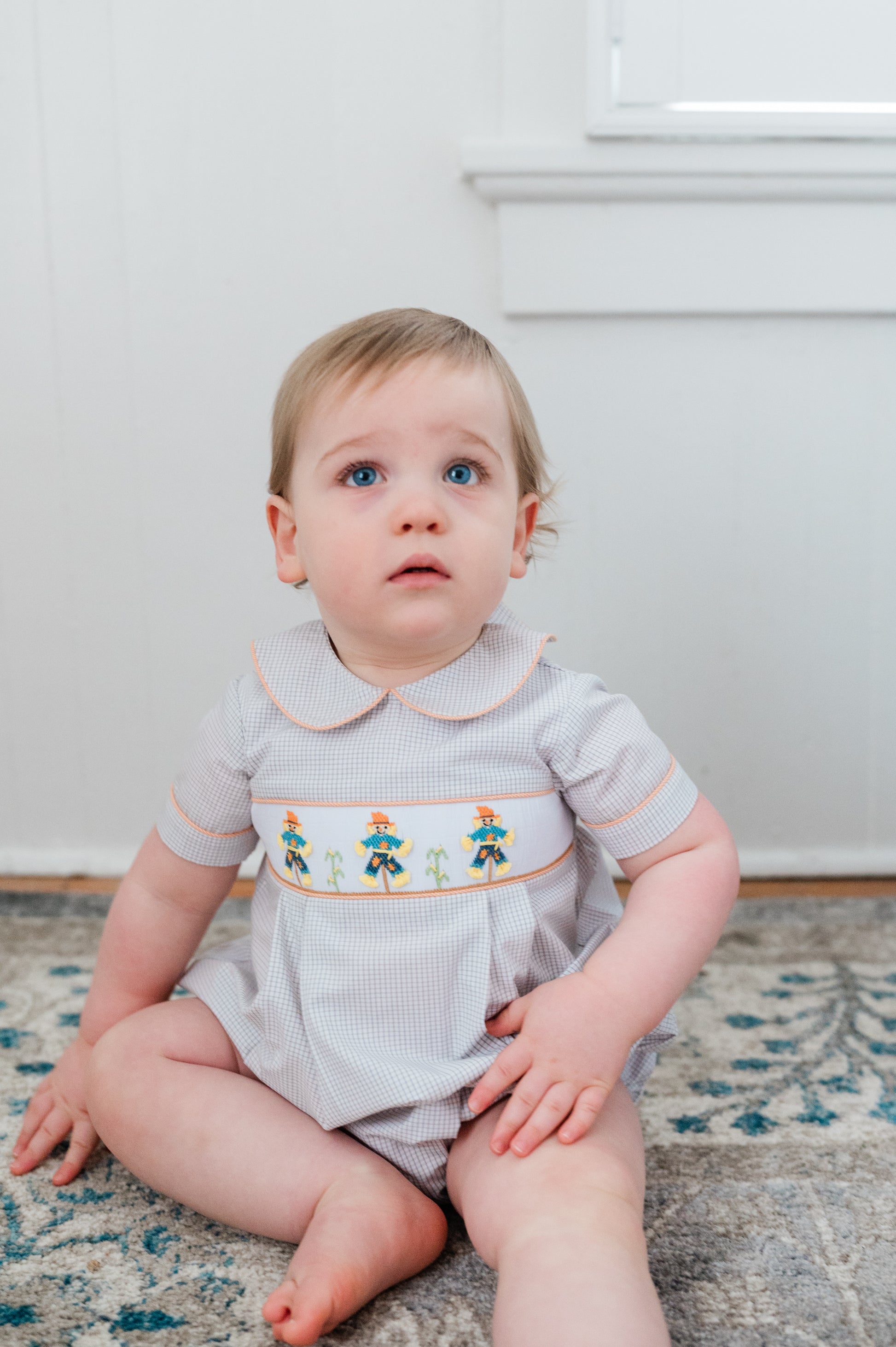 Baby wearing a gray romper with colorful embroidery sitting on a patterned rug.