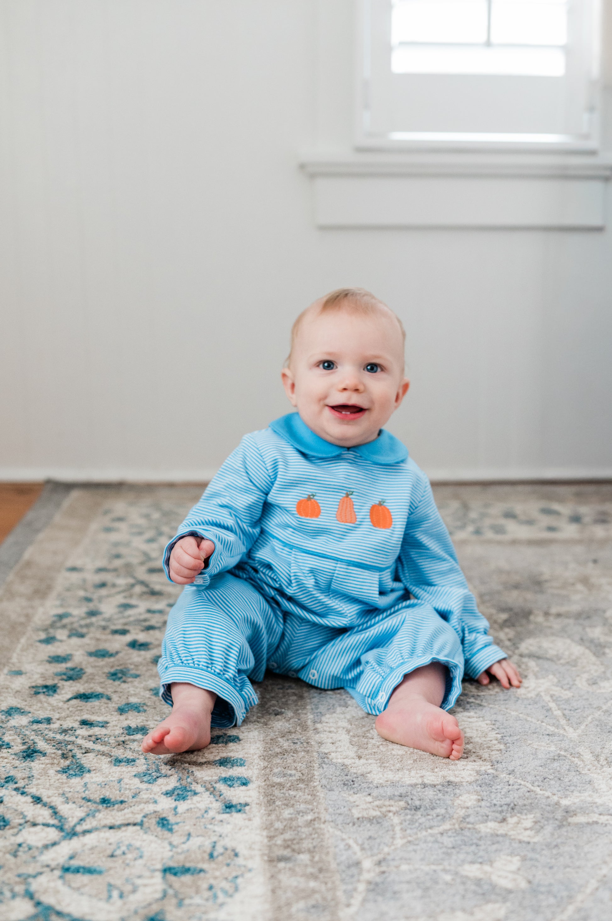 Baby sitting on a rug wearing a blue outfit with orange pumpkins.