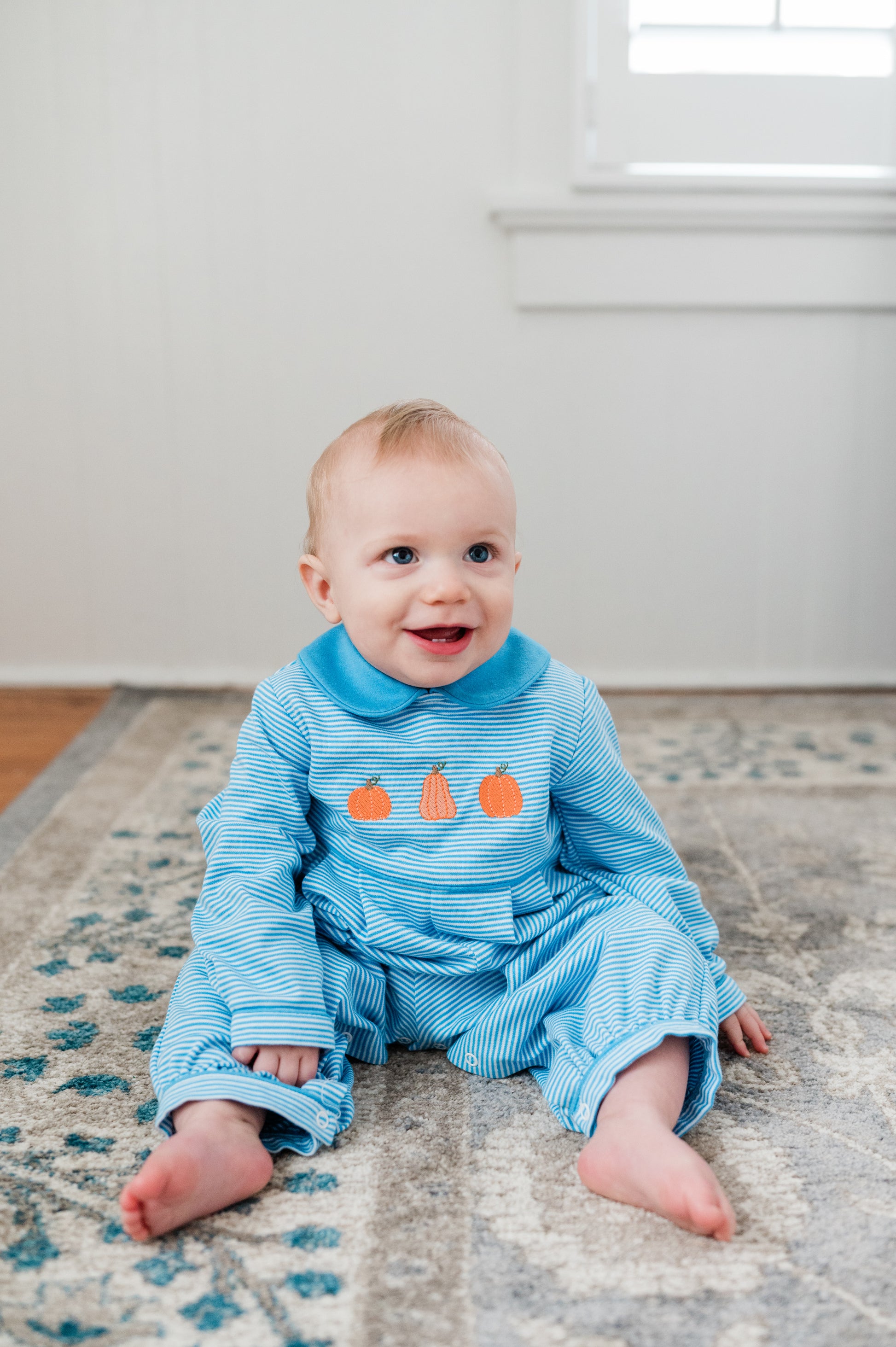 Baby wearing a blue outfit with orange pumpkin design on a patterned rug.