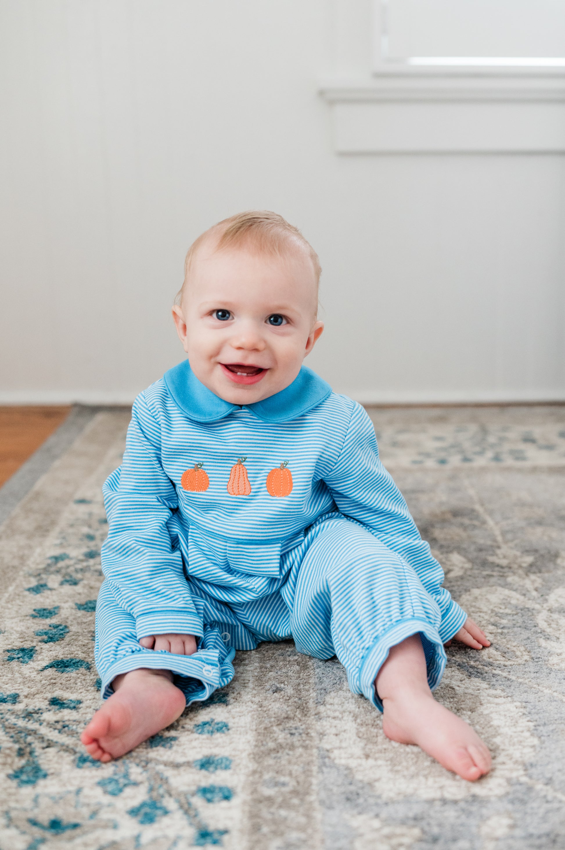 Baby wearing a blue outfit with orange accents sitting on a rug.