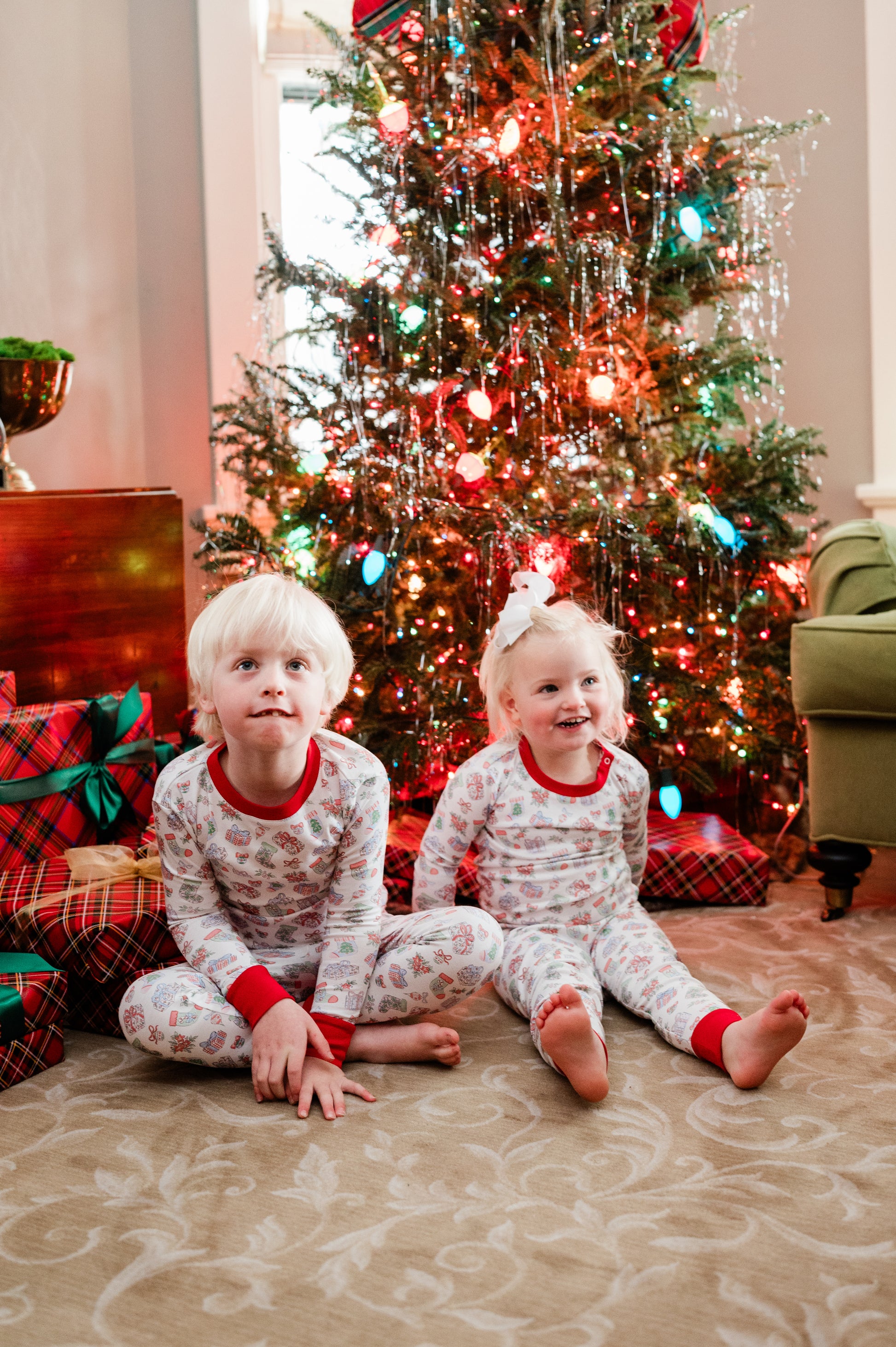 Two children in pajamas sitting on the floor in front of a Christmas tree with presents.