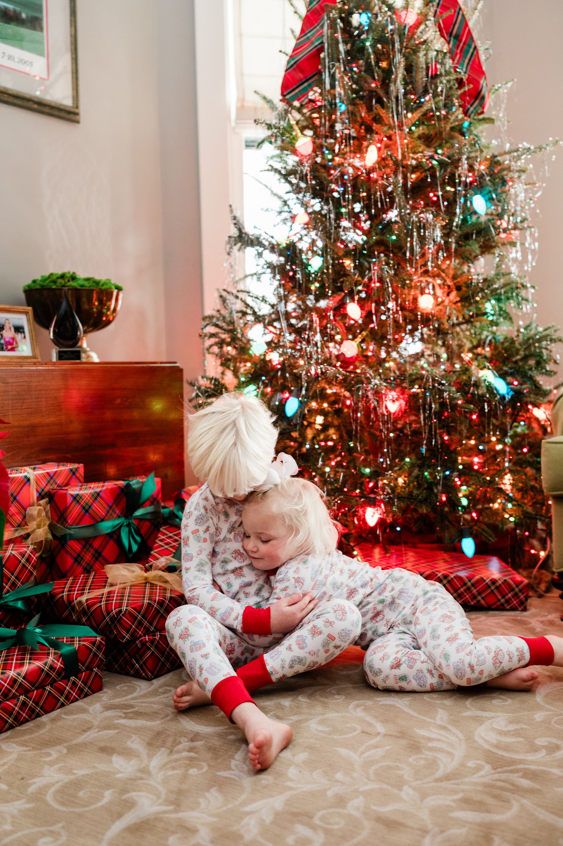 Two children in pajamas sitting on the floor in front of a Christmas tree with presents.