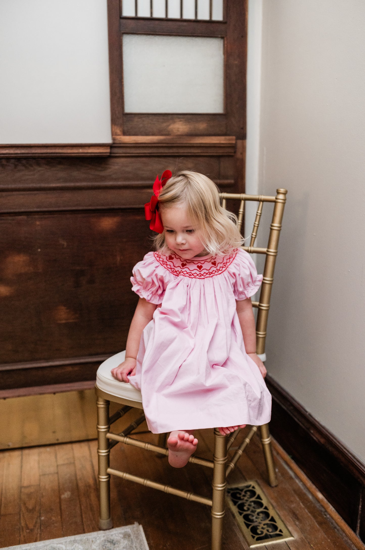 Young girl in a pink dress with red embroidery sitting on a gold chair.