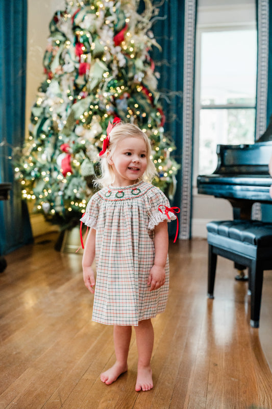 Young girl in a plaid dress standing in front of a decorated Christmas tree in a room with a piano.