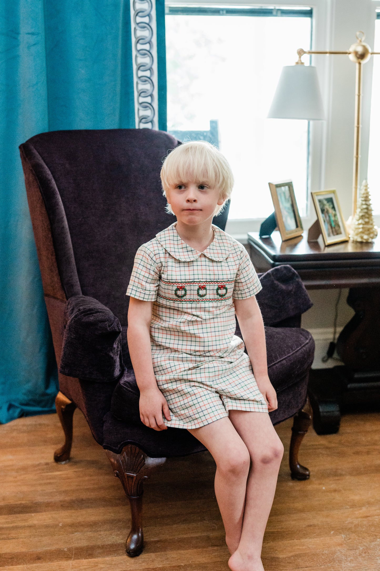 Young boy in a plaid outfit sitting on a chair in a room with a desk and lamp.