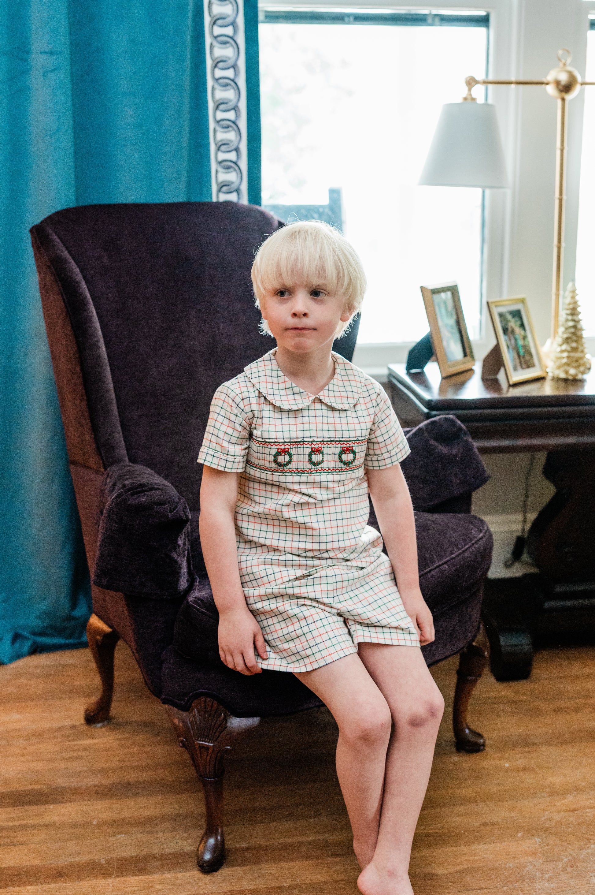 Young boy in a plaid outfit sitting on a chair in a room with a desk and lamp.