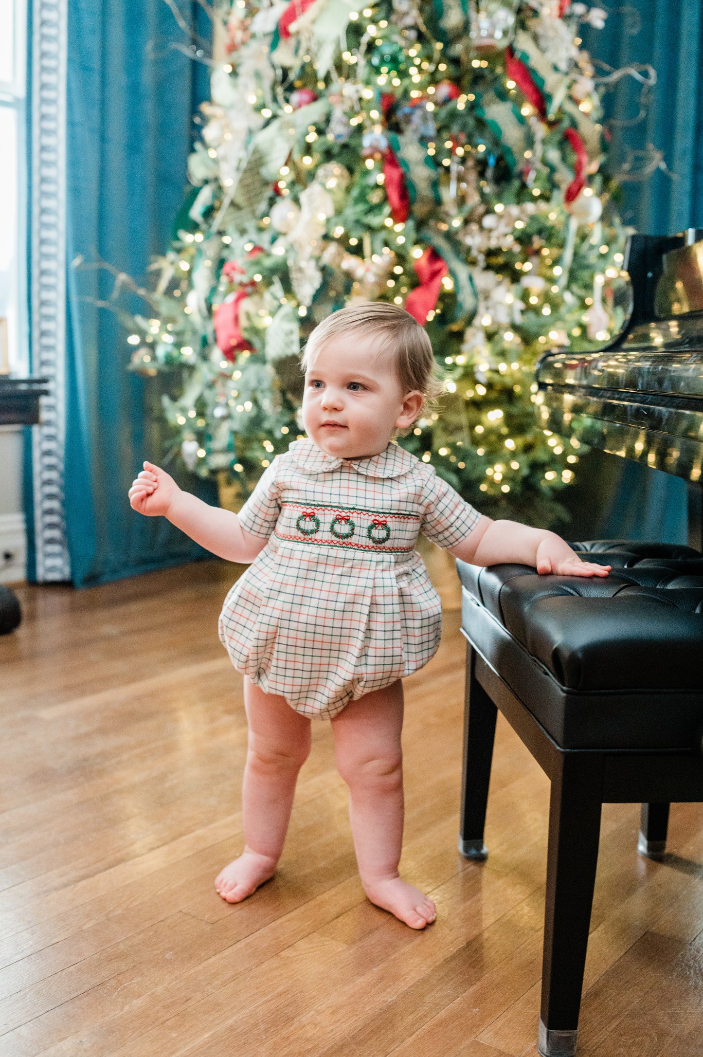 Baby in a festive outfit standing in front of a decorated Christmas tree.