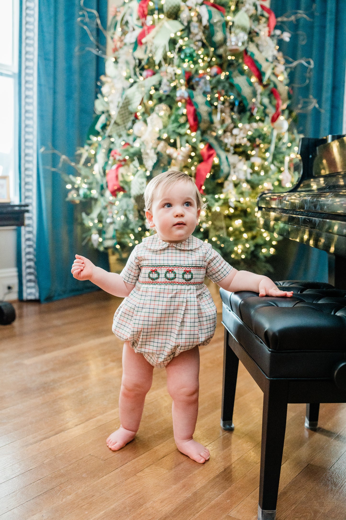 Child in a festive outfit standing in front of a decorated Christmas tree with a piano.