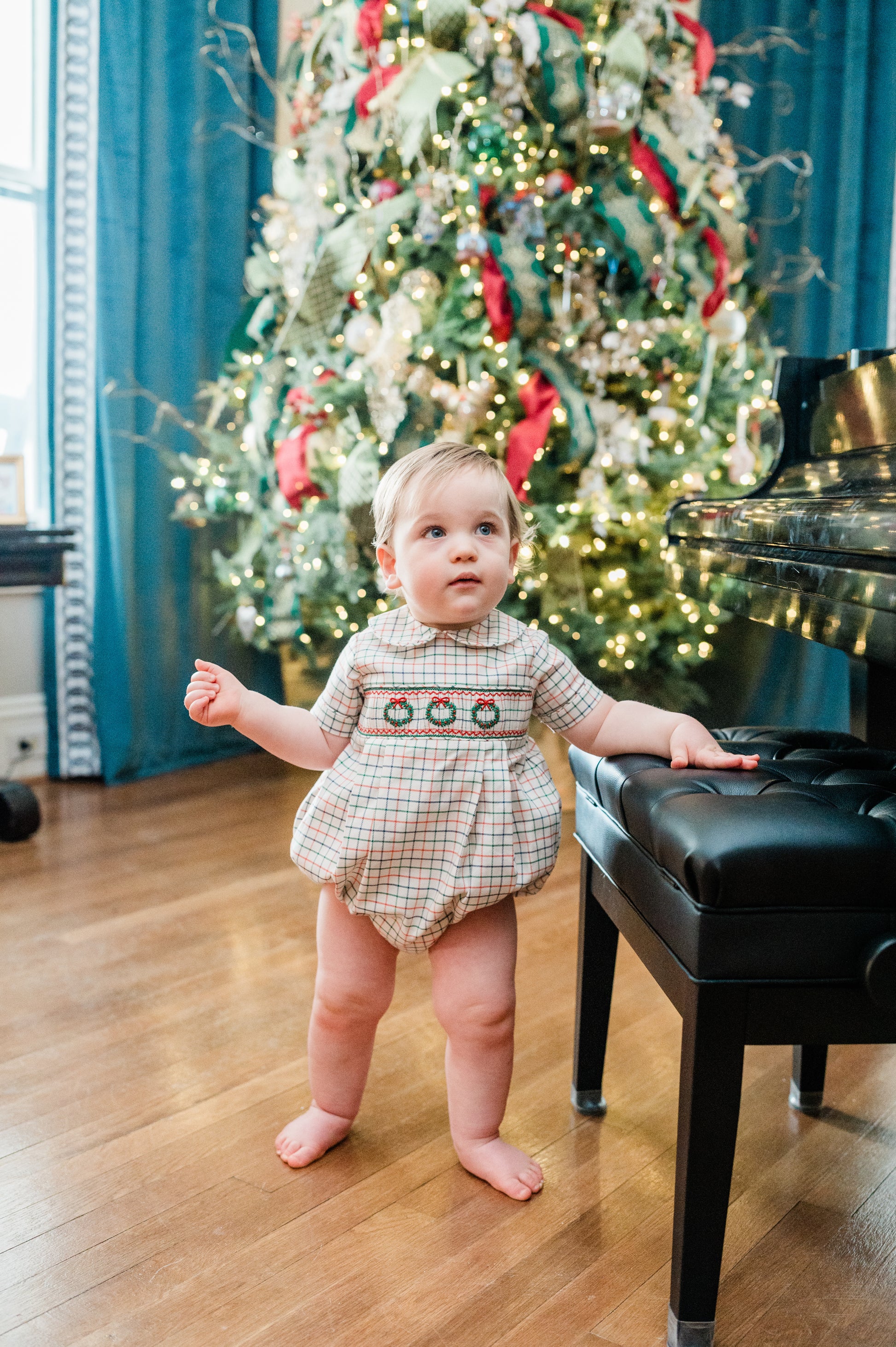Child in a festive outfit standing in front of a decorated Christmas tree with a piano.