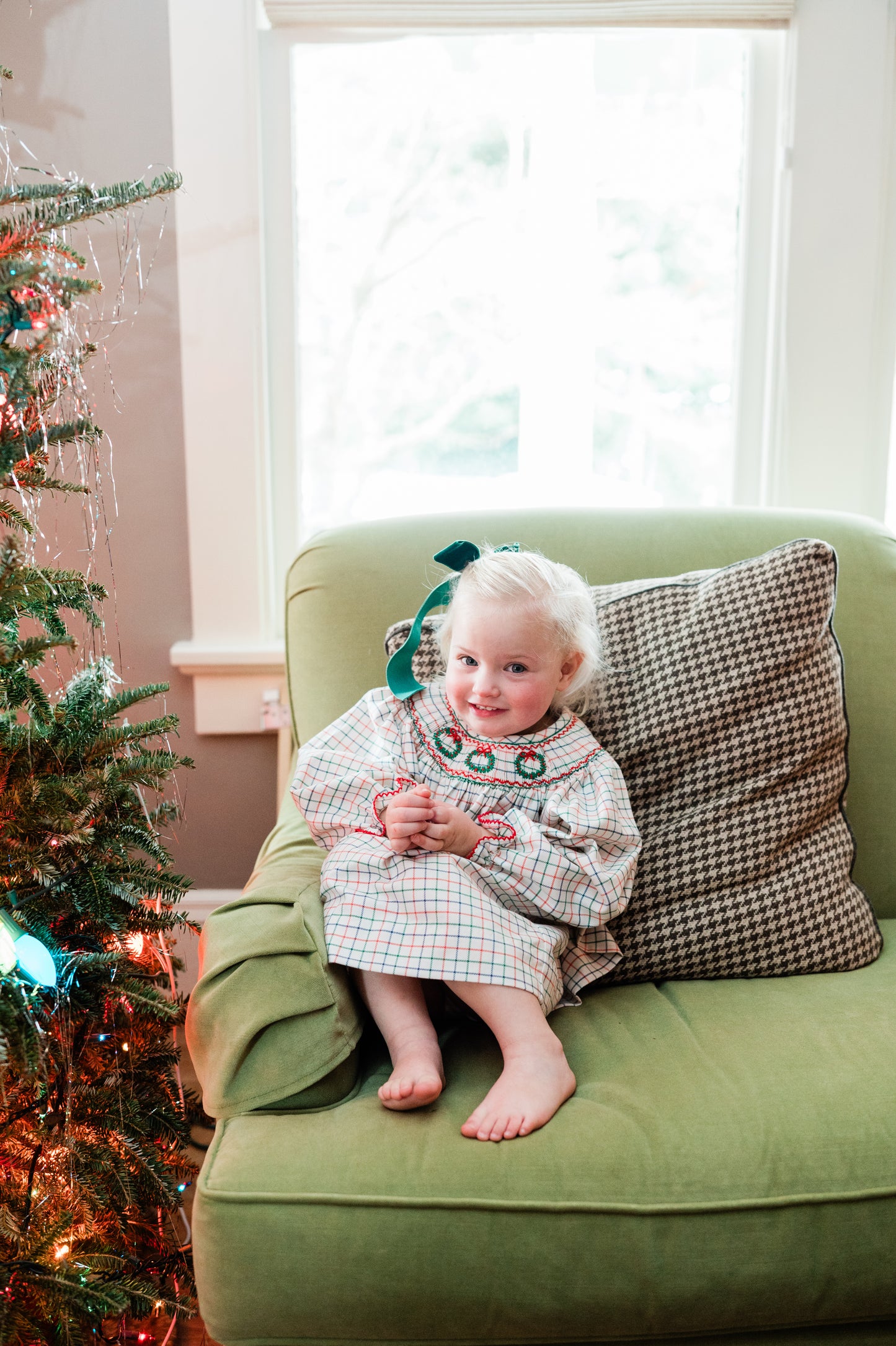 Child in a festive outfit sitting on a green couch next to a decorated Christmas tree.