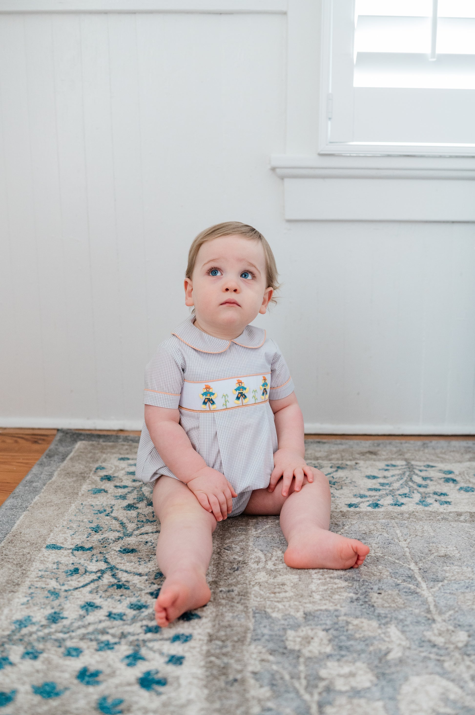 Baby sitting on a patterned rug in a room with white walls and a window.