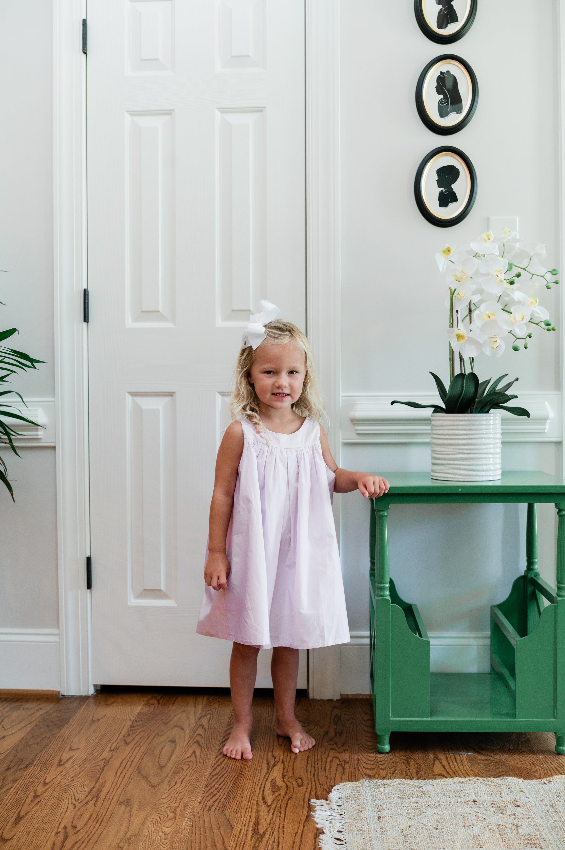 Young girl in a lavender dress standing next to a green table with a white vase and plant in a room.