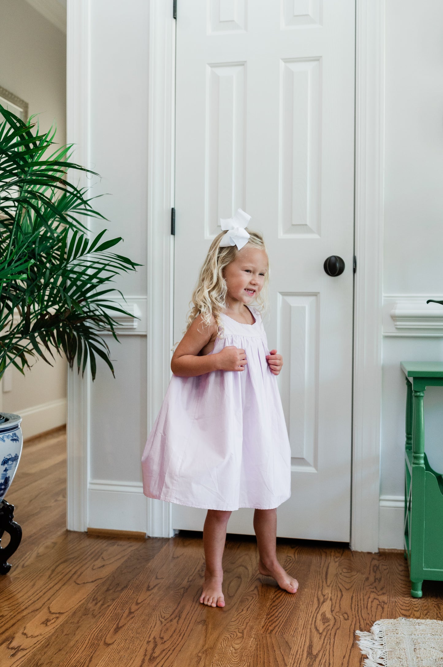 Young girl in a lavender dress standing in a room with wooden floor and white door.