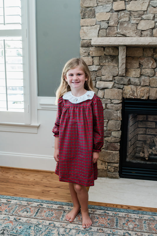 Young girl in a red plaid dress standing in front of a stone fireplace.